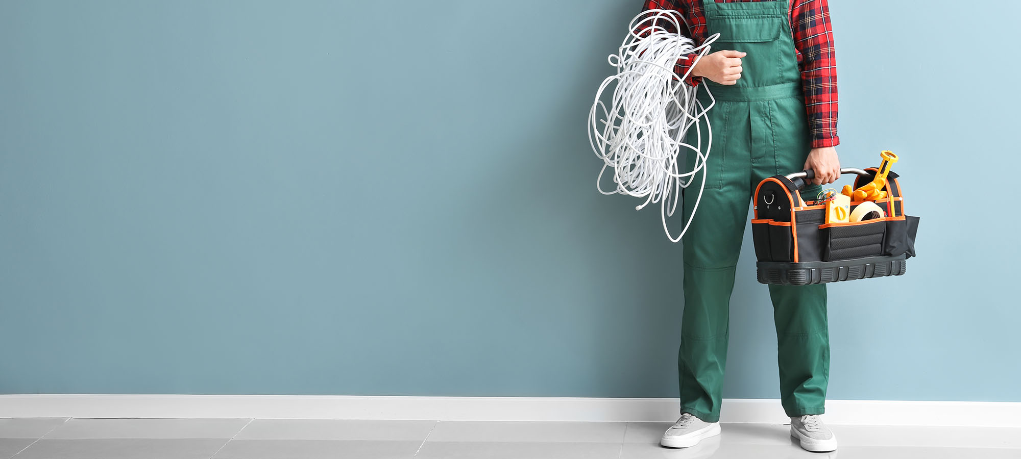 Halbansicht eines Elektrikers mit grünem Overall und rot-karierter Bluse. Er hält in der einen Hand ein aufgewickeltes weißes Kabel und in der anderen eine offene Werkzeugtasche mit Kabelbindern, Isolierband und Werkzeugen, vor hellblauer Wand.
